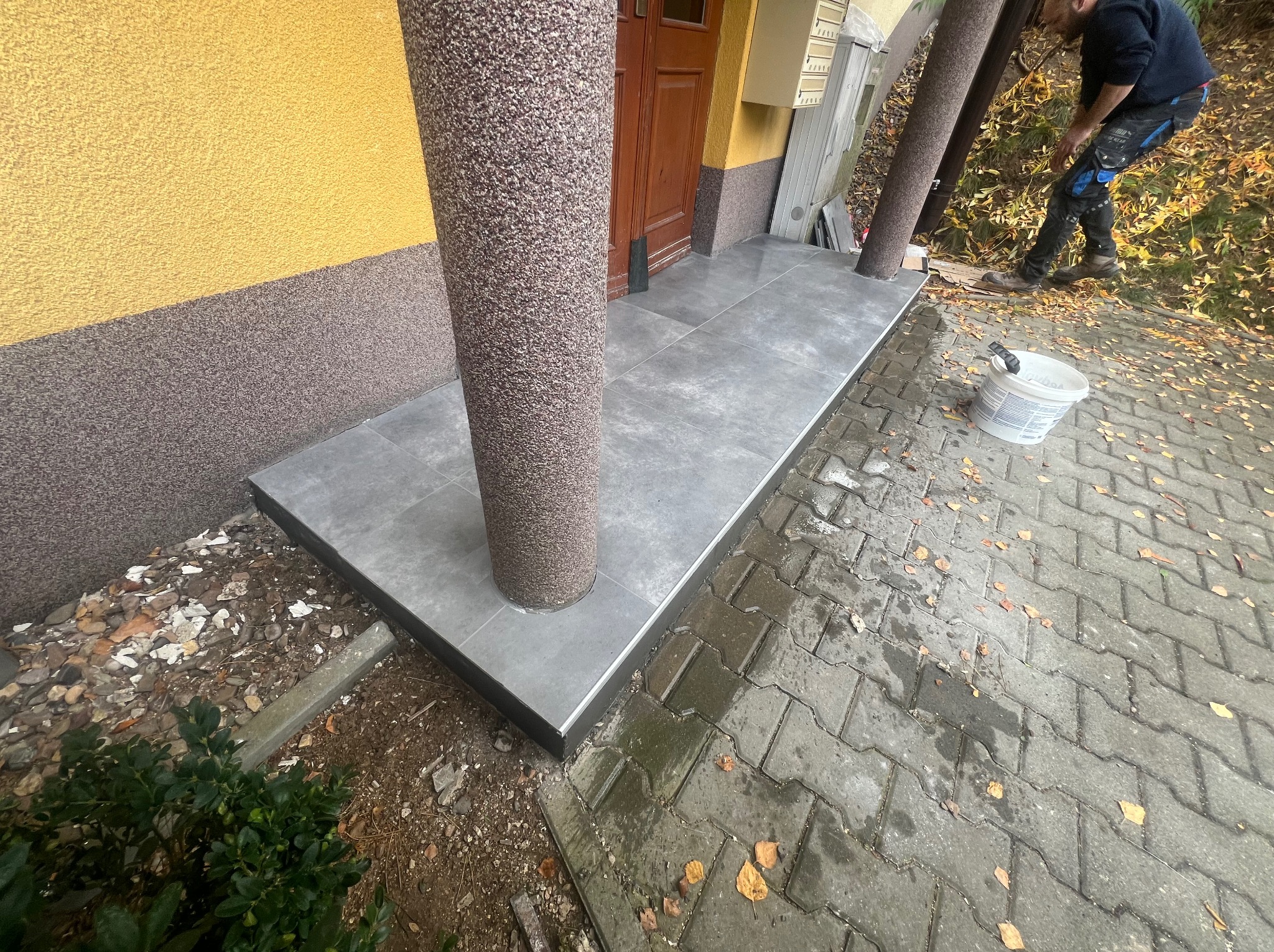 Wykończone płytkami wejście do budynku z kolumnami. Widoczny fragment elewacji i pracownik w tle. Szare płytki z czarnym obramowaniem.