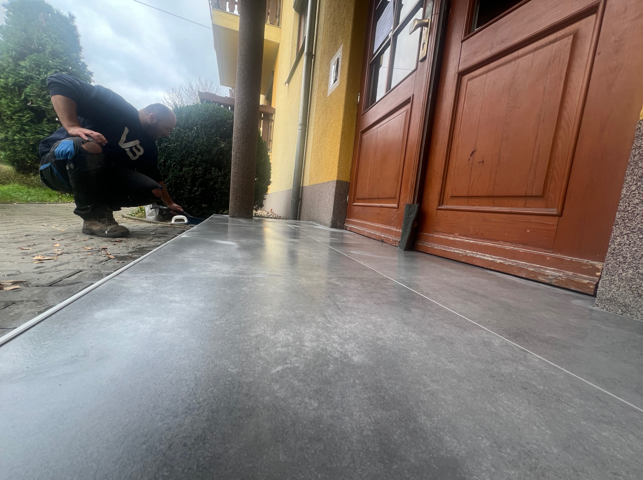 Układanie szarych płytek na schodach wejściowych do domu. Fachowiec w trakcie pracy, widoczne narzędzia i fragment elewacji budynku z drewnianymi drzwiami.