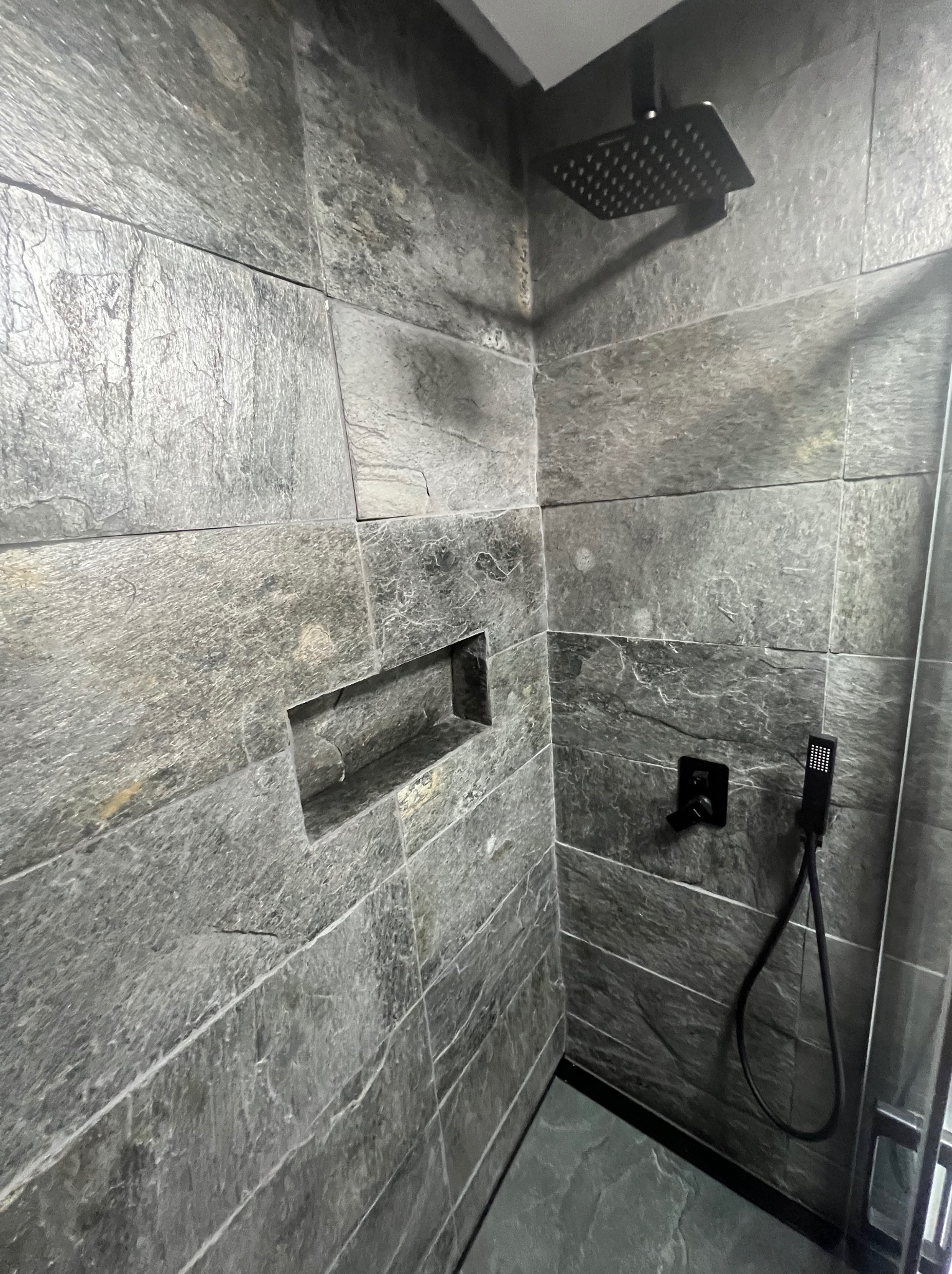 Nowoczesna kabina prysznicowa z szarymi płytkami imitującymi kamień, wnęka na kosmetyki, czarna deszczownica i bateria. Minimalistyczny design.