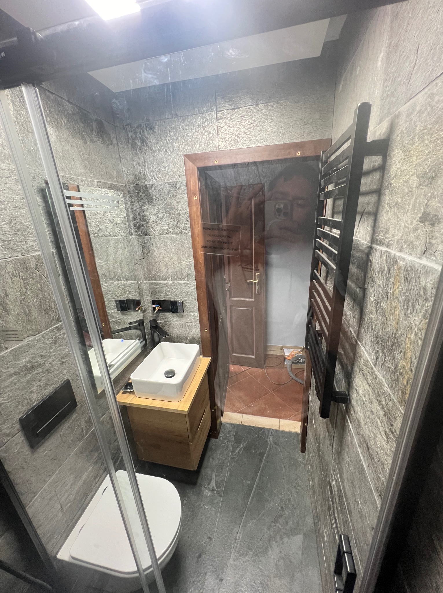 Wykończona łazienka w Pielgrzymce z kabiną prysznicową, umywalką na drewnianej szafce i grzejnikiem drabinkowym na ścianie z szarych płytek imitujących kamień.