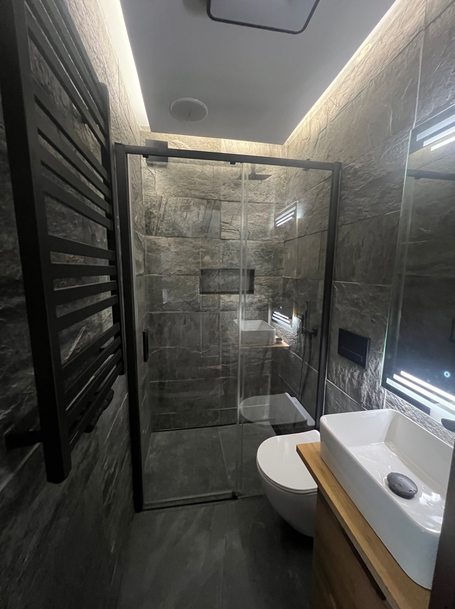 Nowoczesna łazienka z kabiną prysznicową typu walk-in, szarą kamienną okładziną ścian, podświetleniem LED i czarnym grzejnikiem drabinkowym.