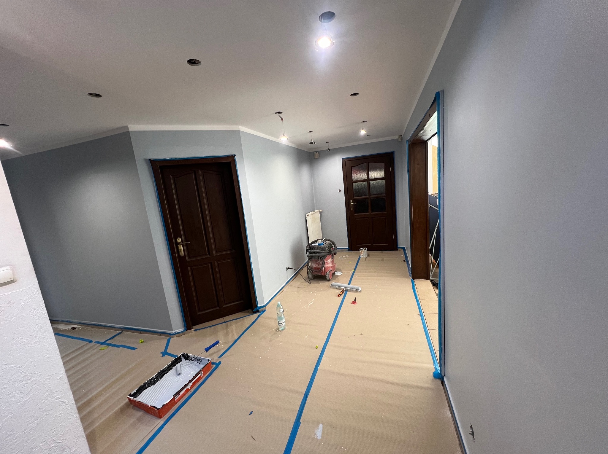 Świeżo pomalowane na szaro ściany w pokoju, zabezpieczone folią i taśmą malarską. Widoczny wałek, kuweta z farbą, odkurzacz i dwie pary drzwi.