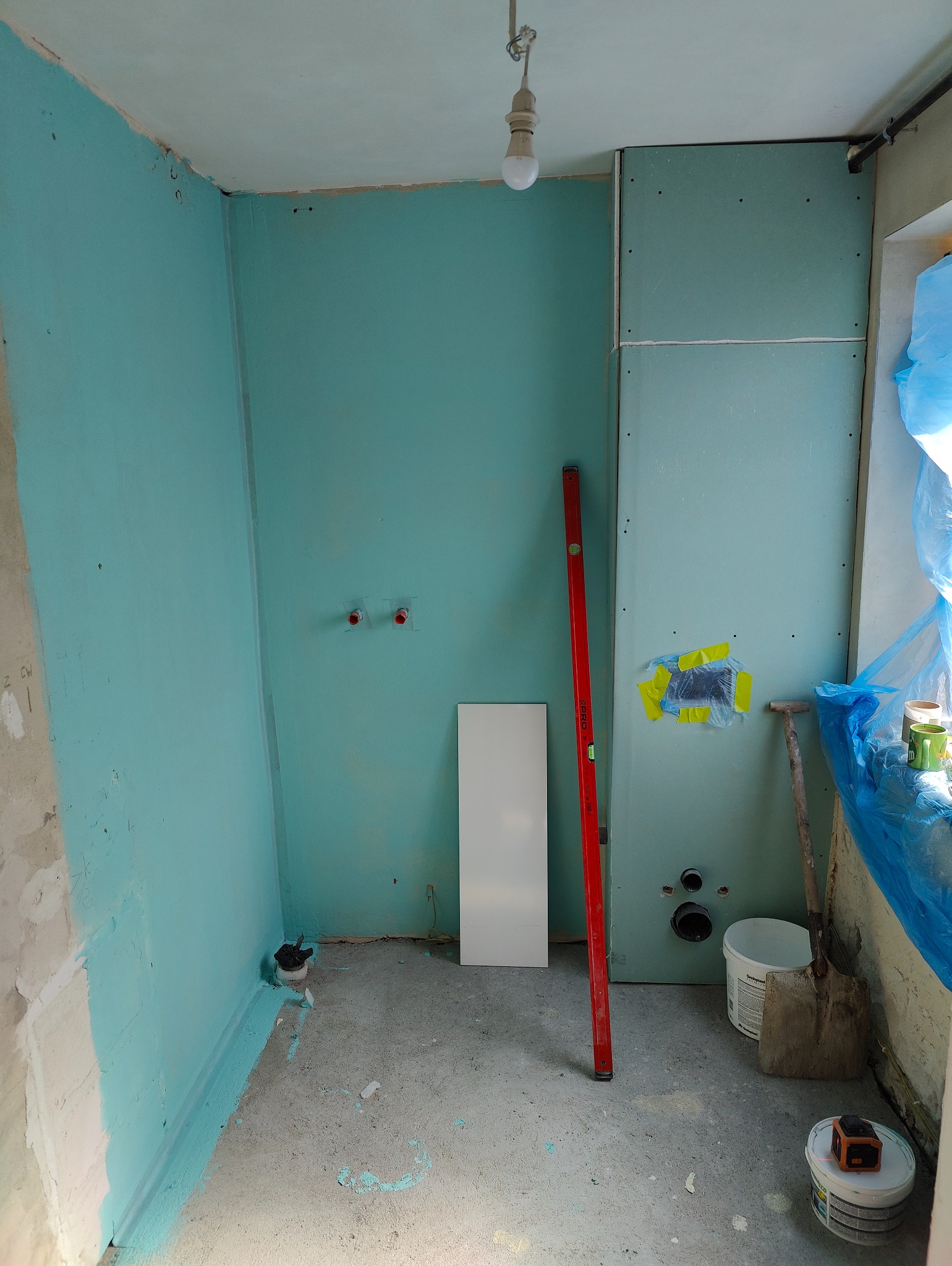 Wnętrze łazienki w trakcie prac: ściany częściowo pokryte turkusową farbą, zamontowane płyty kartonowo-gipsowe, widoczny poziomica, przygotowane przyłącza wodne i kanalizacyjne, oraz narzędzia pracy.
