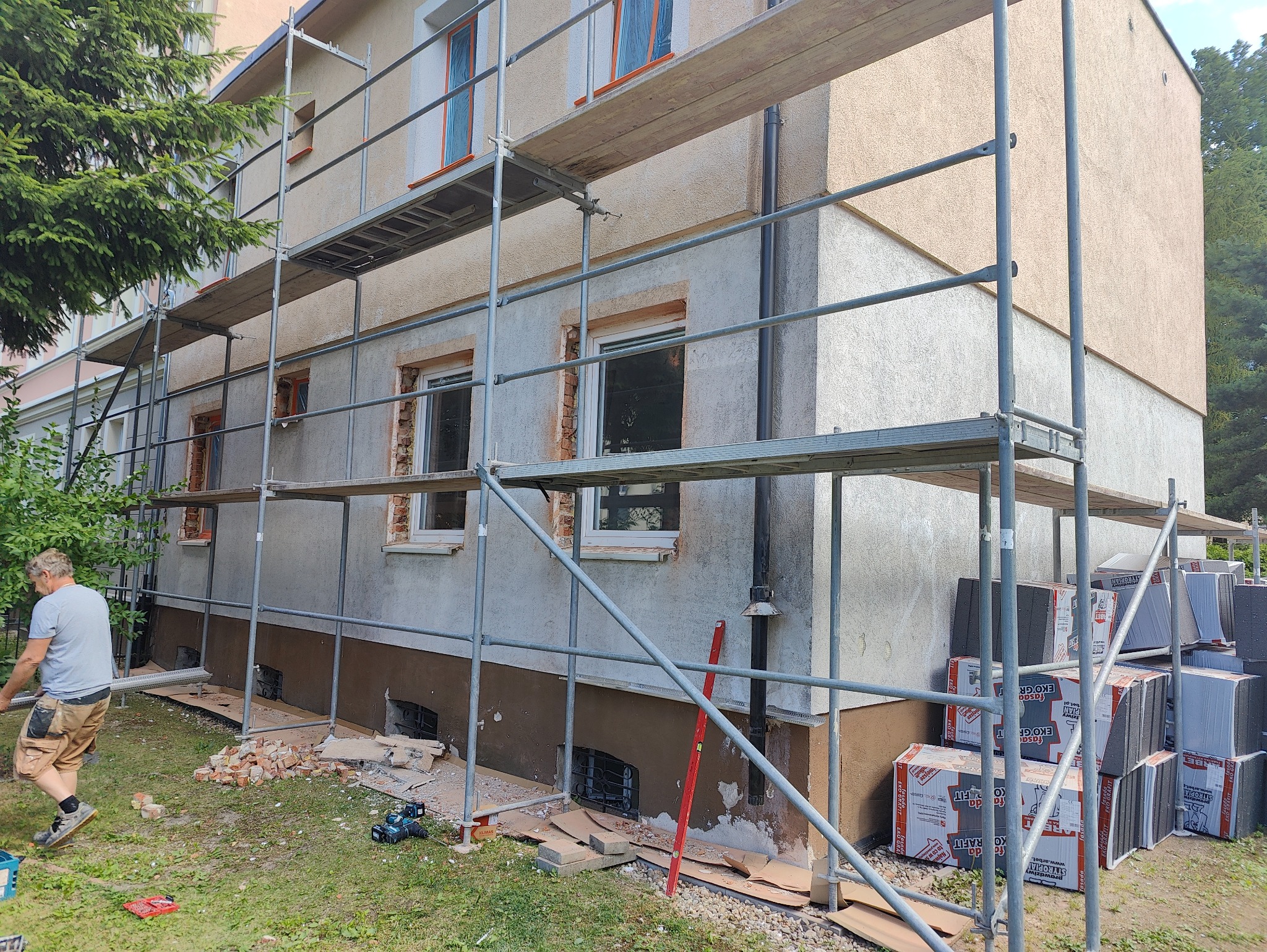 Budynek w trakcie ocieplania, widoczne rusztowania, styropian fasadowy, resztki gruzu i pracownik w tle.
