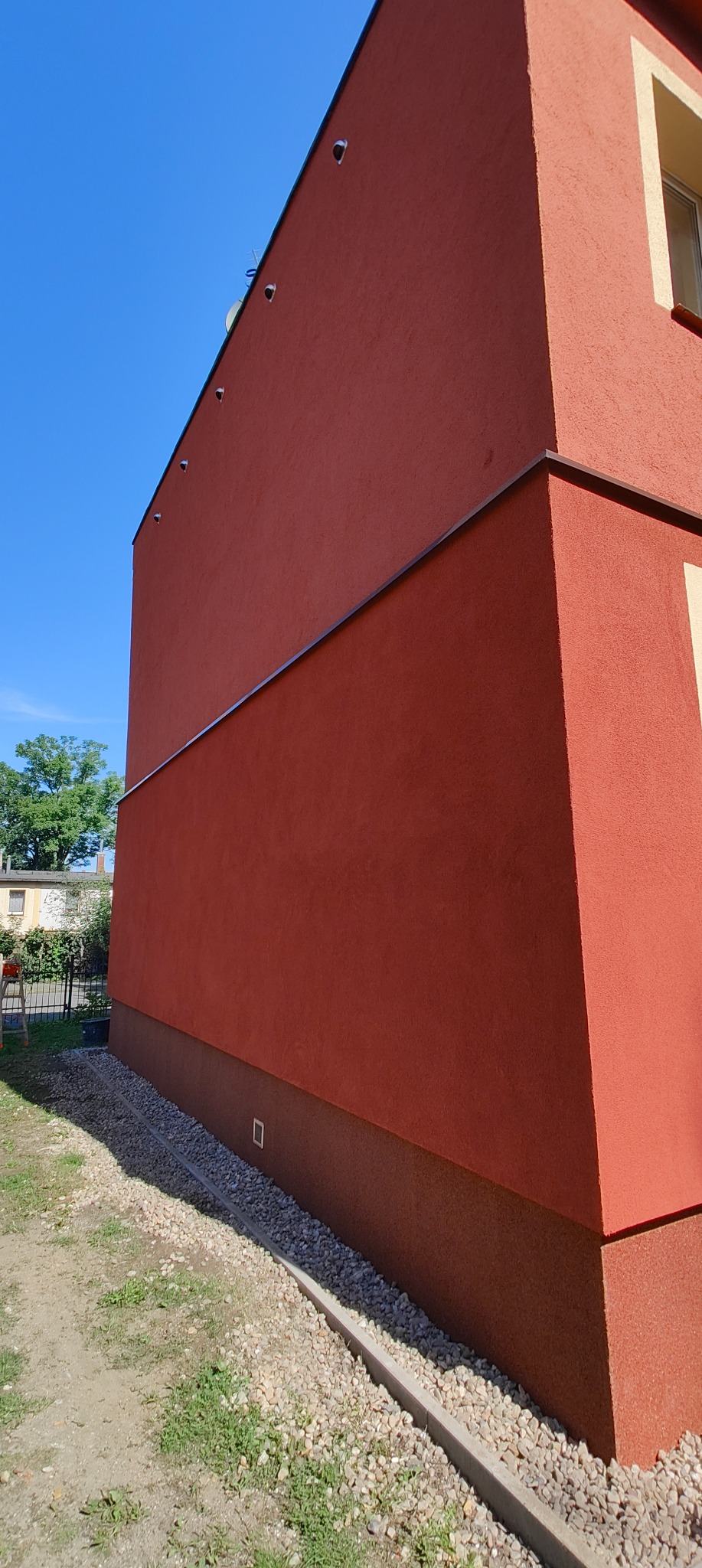 Czerwona ściana budynku z wykończonym tynkiem zewnętrznym, widoczne detale takie jak listwa okapowa, oświetlenie elewacyjne i cokół w innym odcieniu. U podstawy ściany ułożony drenaż z kamieni.