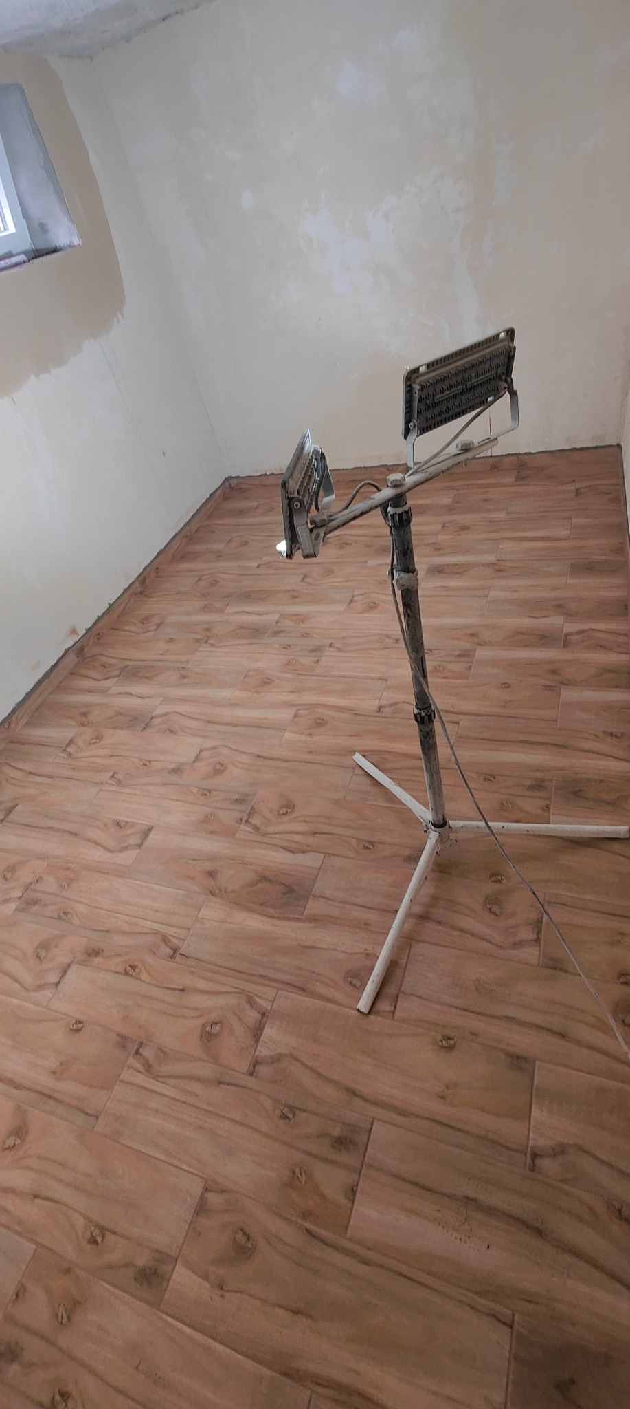 Ukończona podłoga z płytek imitujących drewno w jasnym odcieniu, oświetlona przez lampę budowlaną na trójnogu, ściany w trakcie wykańczania.