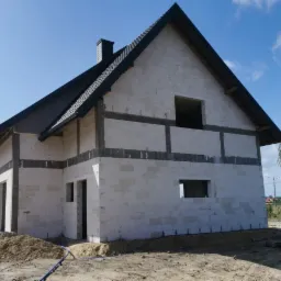 Surowy stan domu z białego bloczka, czarny dach, widoczne otwory okienne i drzwiowe, przygotowany teren pod budowę.