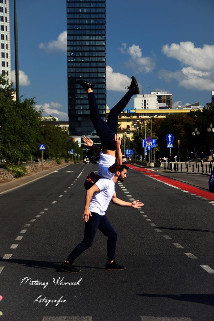 Akrobatyczne ujęcie dwóch mężczyzn na pasach w centrum miasta, jeden stoi na ramionach drugiego w pozycji odwróconej, wysokie budynki w tle.