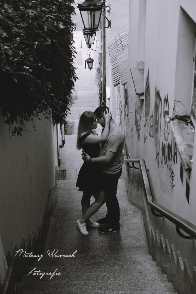 Czarno-białe zdjęcie pary obejmującej się na schodach w wąskiej uliczce z graffiti na ścianach, oświetlonej wiszącymi latarniami.