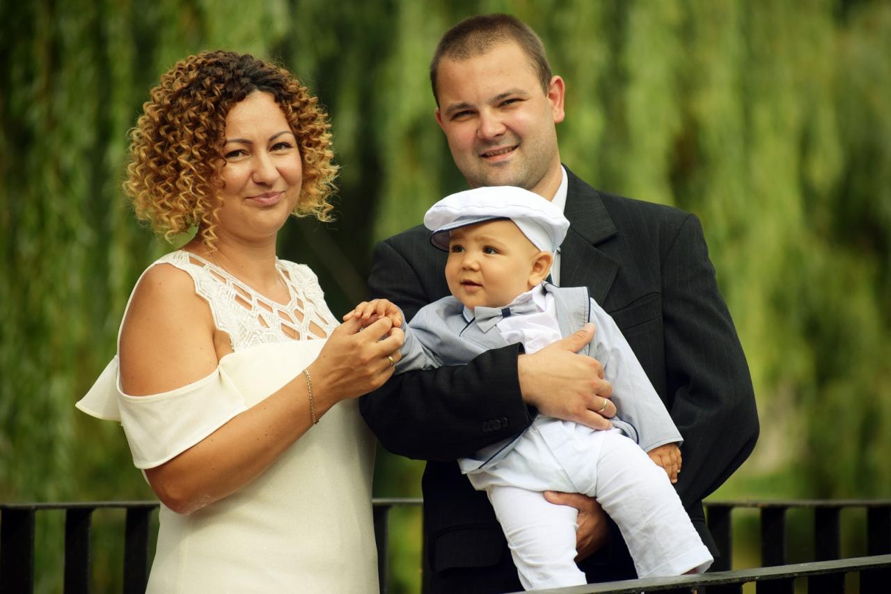 Uśmiechnięta rodzina: mama z kręconymi włosami w białej sukience, tata w garniturze trzymający na rękach elegancko ubranego synka w czapce.