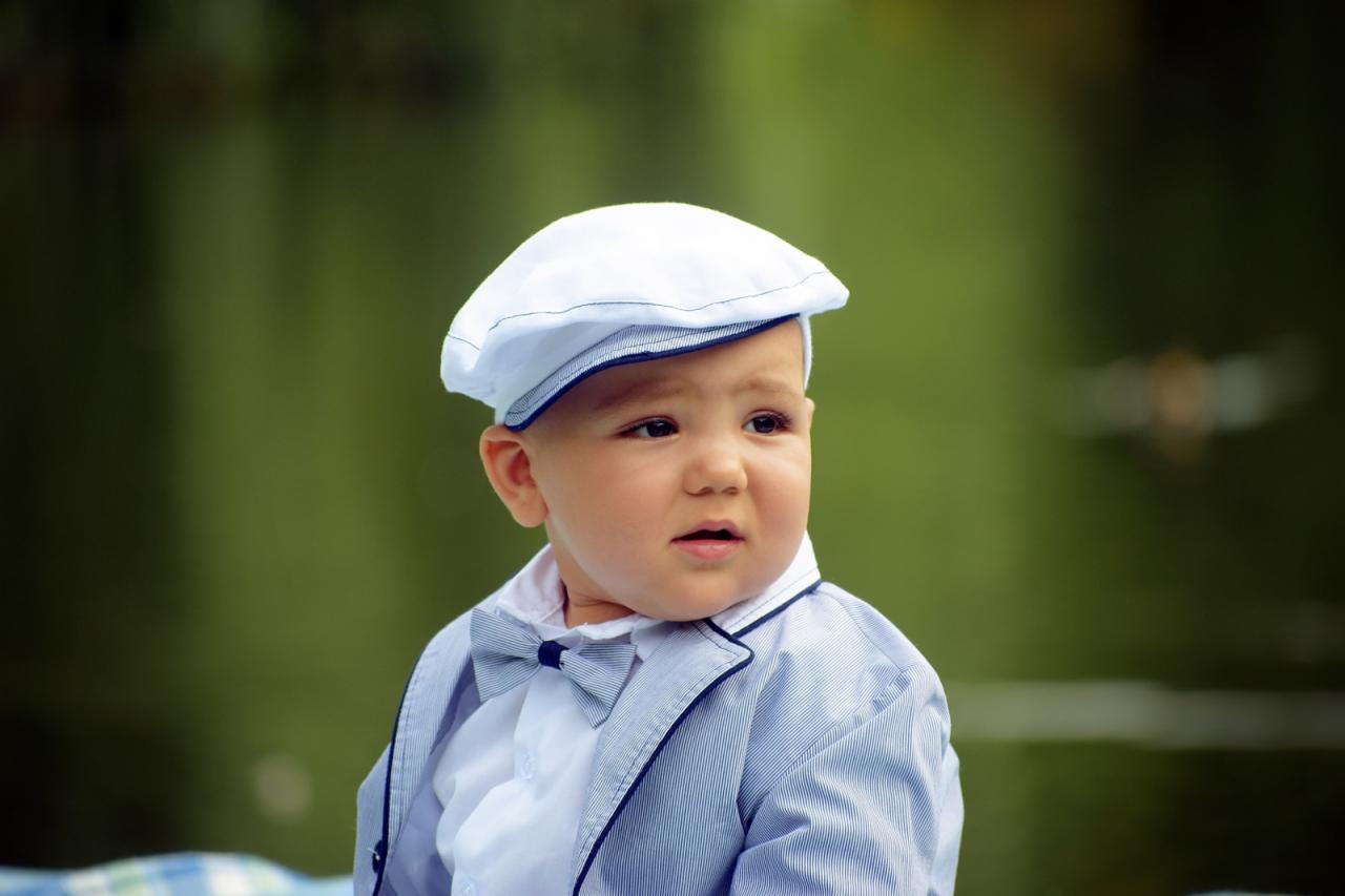 Uroczy chłopiec w eleganckim stroju: biała czapka kaszkietówka, koszula, muszka i marynarka w paski, na tle zieleni.