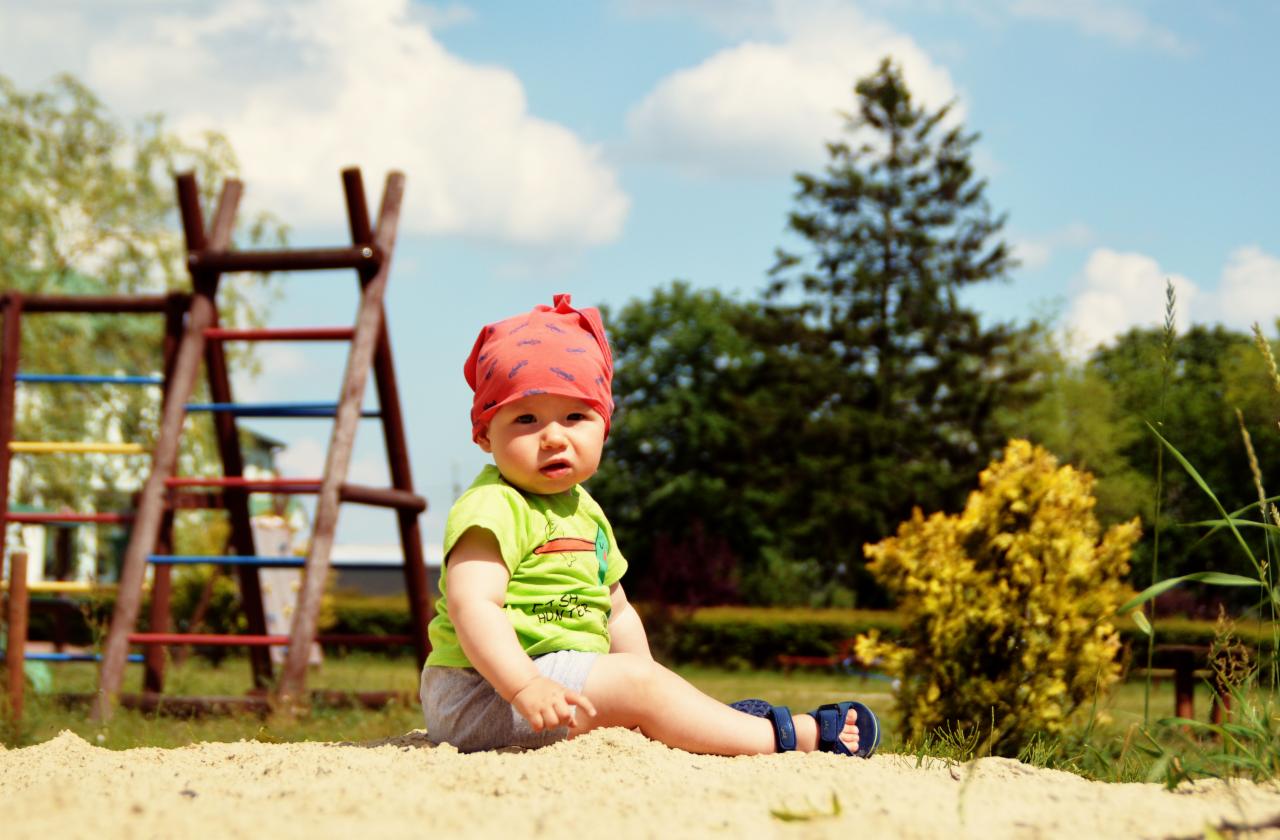 Mały chłopiec w czerwonej chuście na głowie, siedzący w piasku na placu zabaw, z drewnianą drabinką i zielenią w tle.