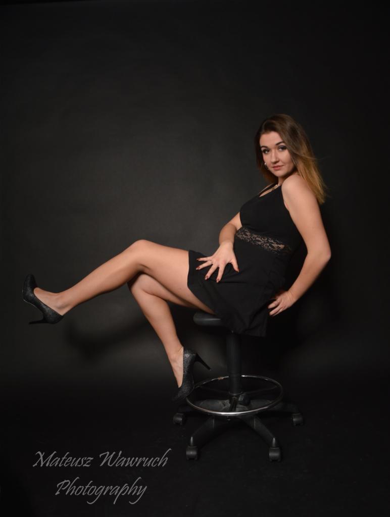 Kobieta w czarnej sukience, siedząca na krześle na czarnym tle, z ręką na brzuchu i nogami uniesionymi, w szpilkach. Widoczny podpis fotografa.