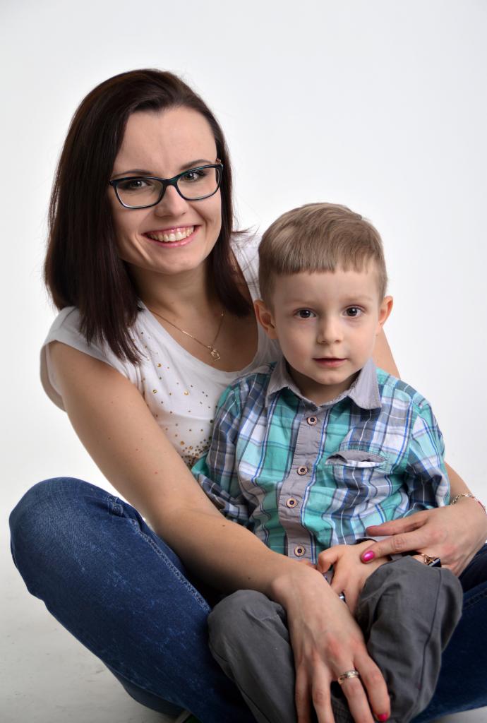 Portret uśmiechniętej matki w okularach, trzymającej na kolanach synka w kraciastej koszuli na jasnym tle.