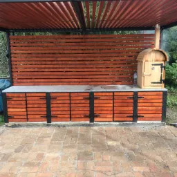 Zadaszona letnia kuchnia z drewnianą zabudową, blatem z szarego kamienia, grillem i piecem wędzarniczym, ustawiona na brukowanym patio.