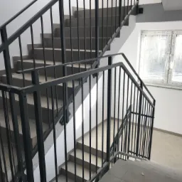 Klatka schodowa wewnątrz budynku z szarymi schodami i czarną metalową balustradą. Widoczne okno z widokiem na drzewa.