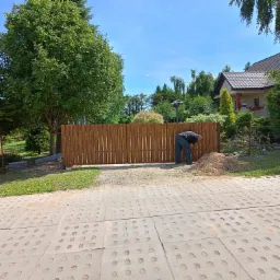 Montaż nowego, drewnianego ogrodzenia panelowego na posesji, widok z zewnątrz na osobę pracującą przy słupku.