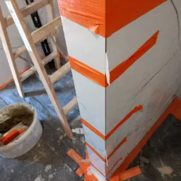 Narożnik obłożony białymi płytkami imitującymi marmur, zabezpieczony pomarańczową taśmą malarską podczas prac wykończeniowych, widoczna drabina i wiadro z zaprawą.