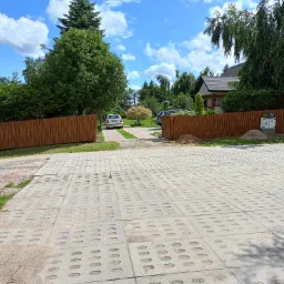 Ułożona nawierzchnia z ażurowych płyt betonowych prowadząca do posesji z drewnianym ogrodzeniem i zielenią w tle.