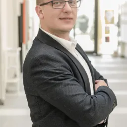 Portret mężczyzny w okularach i ciemnej marynarce, stojącego na jasnym korytarzu z rękami skrzyżowanymi na piersiach.