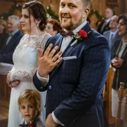 Uśmiechnięty pan młody w granatowym garniturze w kratę, z czerwoną różą w butonierce i bordową muchą, prezentuje obrączkę na palcu podczas ceremonii ślubnej w kościele, obok stoi panna młoda...