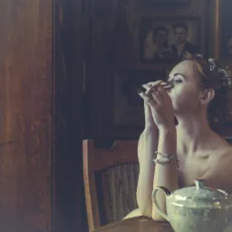 Kobieta z upiętymi włosami i delikatną biżuterią, siedząca przy stole z porcelanowym dzbankiem do herbaty, w tle portrety w ozdobnych ramach i drewniane meble w stylu retro, tworzące intymną...