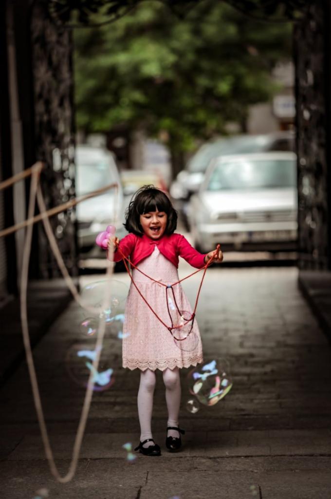 Uśmiechnięta dziewczynka w różowej sukience i czerwonym sweterku puszcza bańki mydlane na tle kamiennej uliczki z samochodami w oddali.