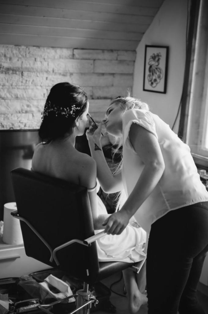 Czarno-białe zdjęcie kobiety w trakcie robienia makijażu przez wizażystkę przed ślubem, widoczna dekoracja we włosach panny młodej i tatuaż na ręce wizażystki.