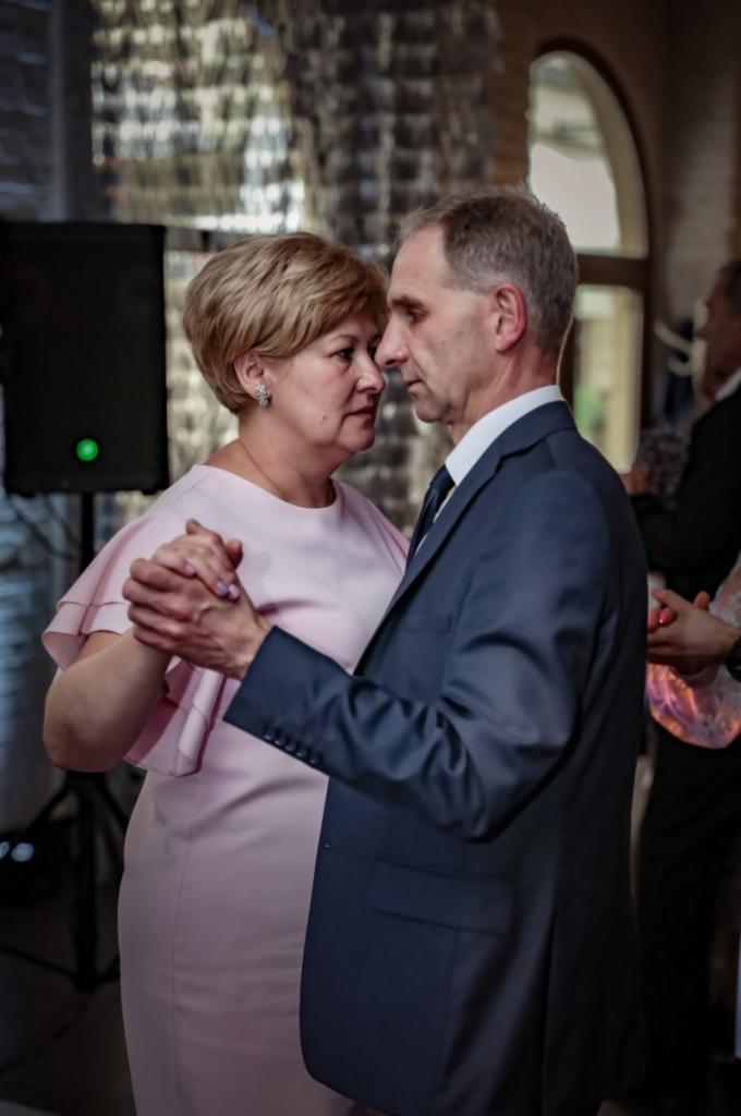 Para w średnim wieku tańczy blisko siebie, kobieta w różowej sukience z falbankami na ramionach i mężczyzna w granatowym garniturze, uchwyceni podczas spokojnego tańca.