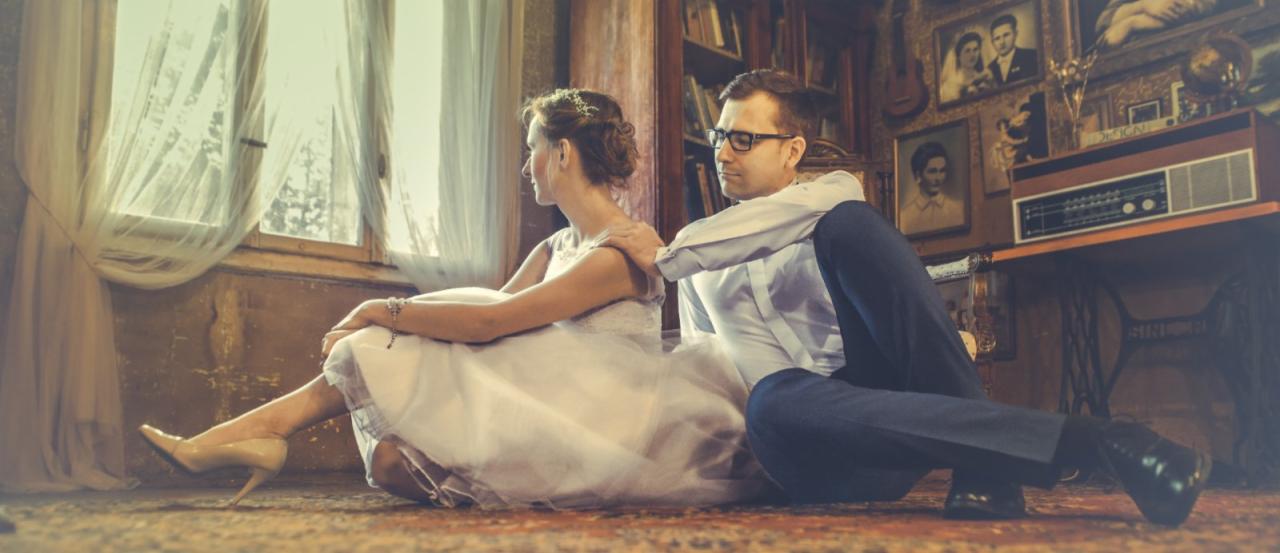 Para młoda siedząca na podłodze w pokoju w stylu retro, ona w białej sukni i beżowych szpilkach, on w koszuli i spodniach, obok okna z firanką, w tle regał z książkami i zdjęcia na ścianie, w tym...