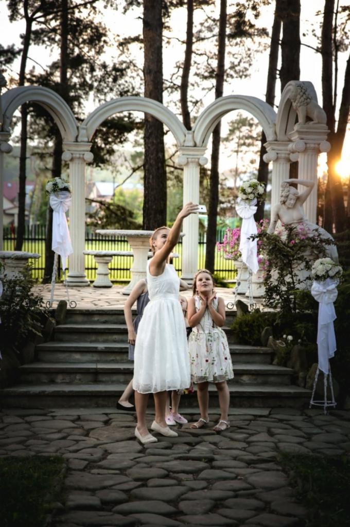 Kobieta w białej sukience robi selfie z dwiema dziewczynkami na tle dekoracji ślubnych: kolumn, łuków, kwiatów i rzeźb w plenerze.
