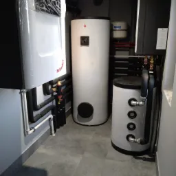 Kotłownia z piecem Zehnder w folii ochronnej, obok dwa białe zasobniki wody, szare rury i płytki podłogowe