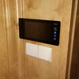 Zamontowany, czarny wideodomofon firmy Orno na drewnianej ścianie obok dwóch białych włączników światła.