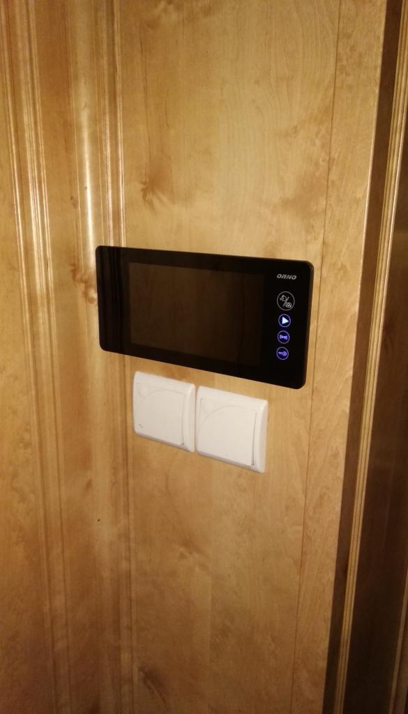 Zamontowany, czarny wideodomofon firmy Orno na drewnianej ścianie obok dwóch białych włączników światła.