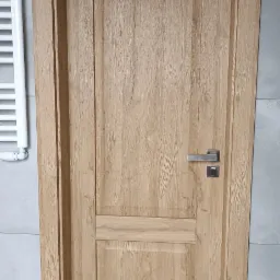 Jasne, drewniane drzwi wewnętrzne z metalową klamką na tle szarej ściany i białego grzejnika drabinkowego. Widoczna struktura drewna i dwuczęściowa konstrukcja skrzydła.