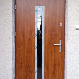 Berlin-Technik Łukasz Berliński - Brązowe drzwi wejściowe z długim, wąskim przeszkleniem, zabezpieczonym folią. Widoczny fragment elewacji i oświetlenie zewnętrzne.