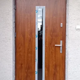 Brązowe drzwi wejściowe z długim, wąskim przeszkleniem, zabezpieczonym folią. Widoczny fragment elewacji i oświetlenie zewnętrzne.