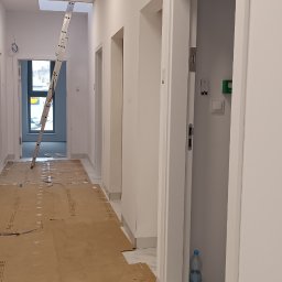 Berlin-Technik Łukasz Berliński - Korytarz w trakcie remontu z zabezpieczoną podłogą kartonem i widoczną drabiną opartą o sufit. Białe ściany i ościeżnice drzwiowe.