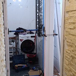 Berlin-Technik Łukasz Berliński - Montaż ościeżnicy drzwiowej w łazience z użyciem poziomicy laserowej. Widoczne narzędzia i przymocowana ościeżnica. Ściana wykończona kamieniem dekoracyjnym.