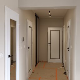 Berlin-Technik Łukasz Berliński - Korytarz w trakcie remontu z widocznymi białymi drzwiami z czarnymi klamkami i zabezpieczoną podłogą. Widoczne lampy sufitowe.