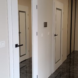 Berlin-Technik Łukasz Berliński - Białe, przeszklone drzwi wewnętrzne z czarną klamką, widoczne w korytarzu z marmurową podłogą. W tle kolejne drzwi i przesuwne drzwi szafy.