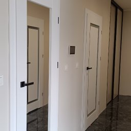 Berlin-Technik Łukasz Berliński - Nowoczesne, białe drzwi wewnętrzne z czarnymi klamkami, widoczne w perspektywie korytarza z marmurową podłogą. Minimalistyczny design, jasne ściany.