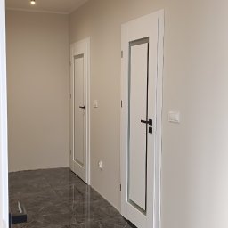 Berlin-Technik Łukasz Berliński - Nowoczesne, białe drzwi wewnętrzne z czarnymi klamkami w korytarzu z ciemną, marmurową podłogą. Minimalistyczny design, punktowe oświetlenie sufitowe.