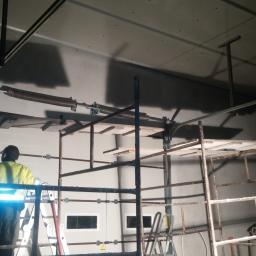 Berlin-Technik Łukasz Berliński - Instalacja sprężyny bramy garażowej: monter w żółtej kamizelce na rusztowaniu, widoczny mechanizm sprężynowy i częściowo zamontowana brama segmentowa w surowym wnętrzu garażu.