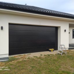 Berlin-Technik Łukasz Berliński - Czarna brama garażowa segmentowa w nowoczesnym domu z białymi ścianami, widoczna drabina i skrzynka narzędziowa na zewnątrz.