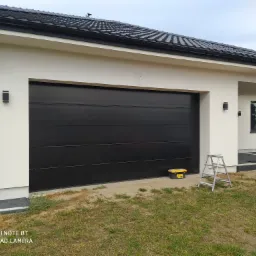 Czarna brama garażowa segmentowa w nowoczesnym domu z białymi ścianami, widoczna drabina i skrzynka narzędziowa na zewnątrz.