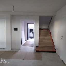 Berlin-Technik Łukasz Berliński - Wnętrze domu w trakcie remontu: widoczne białe drzwi wewnętrzne, schody zabezpieczone kartonem i folią, oraz widok na podjazd z zaparkowanym busem.