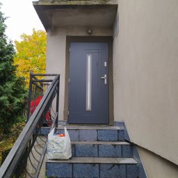 Berlin-Technik Łukasz Berliński - Szare drzwi wejściowe z pionowym przeszkleniem, zamontowane na zewnątrz budynku, widoczne schody wejściowe wyłożone niebieską glazurą, obok torba z zakupami i doniczka z małą roślinką.