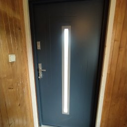 Berlin-Technik Łukasz Berliński - Ciemnoszare drzwi wejściowe z podłużnym przeszkleniem, widoczne od wewnątrz, z drewnianą boazerią i jasną ościeżnicą.