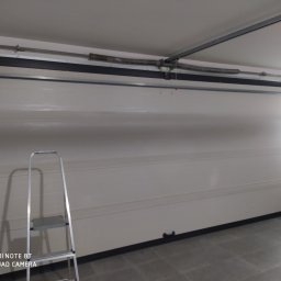 Berlin-Technik Łukasz Berliński - Wnętrze garażu z białą segmentową bramą garażową w pozycji zamkniętej, widoczna drabina aluminiowa i sprężyna nad bramą.